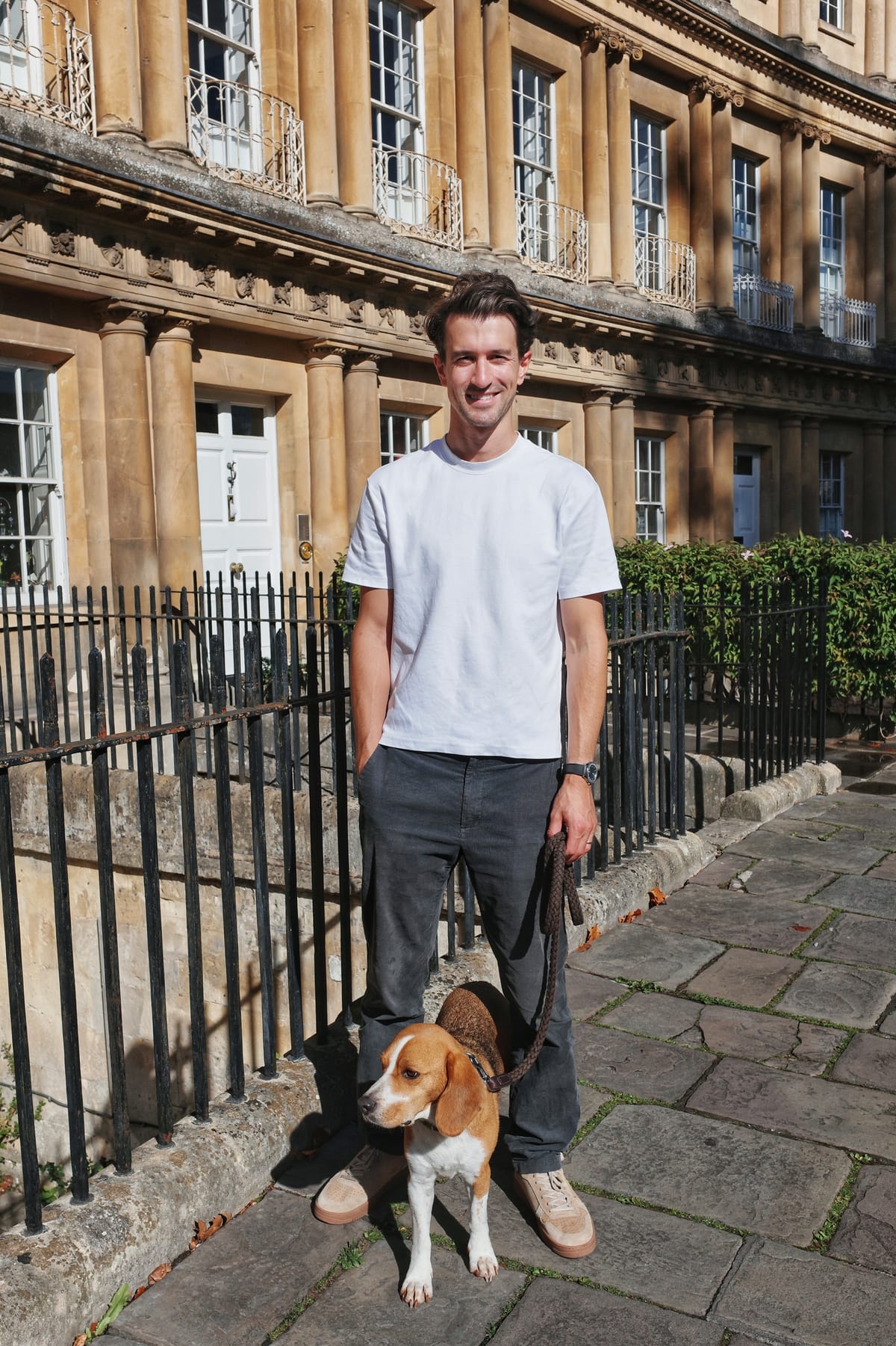 George Pym in a white T-shirt, standing on a stone pavement outside a Georgian terrace with his beagle, Hugo, on a lead.